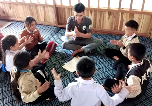 Six Lao children sit on the floor, learning Lao sign language from a young teacher.