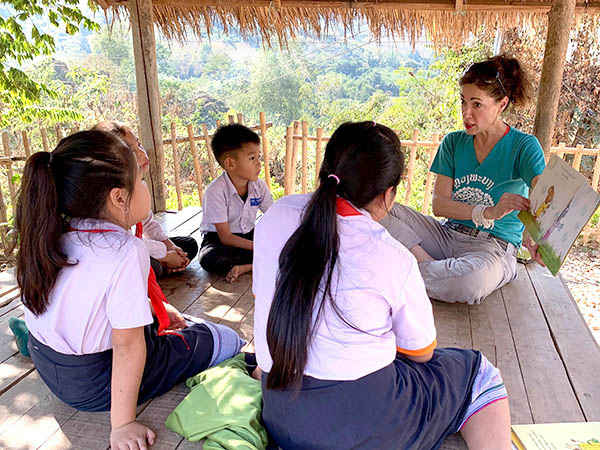 A woman reads aloud and shows pictures in a book to four Lao schoolchildren, all sitting in a small hut